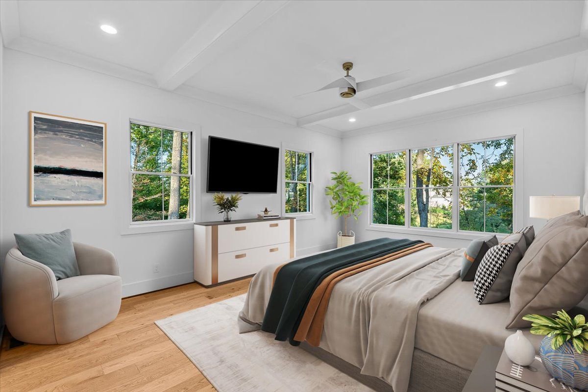 Bright bedroom with a large bed, windows, TV, and armchair. Sunlight streams in.