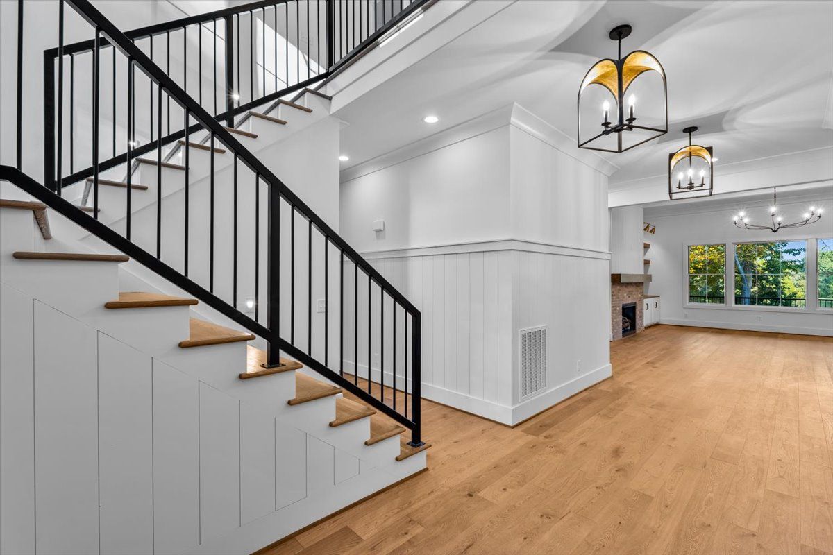 Staircase with black railing and white walls, leading up. Hardwood floors and gold chandeliers in the background.