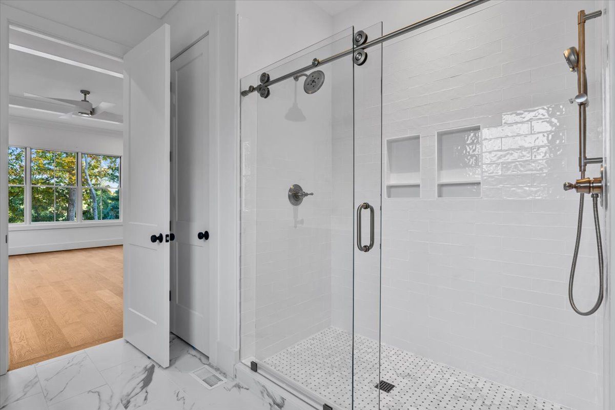 Bright, modern bathroom with glass shower, white tile, and a doorway to a bedroom with hardwood floors.