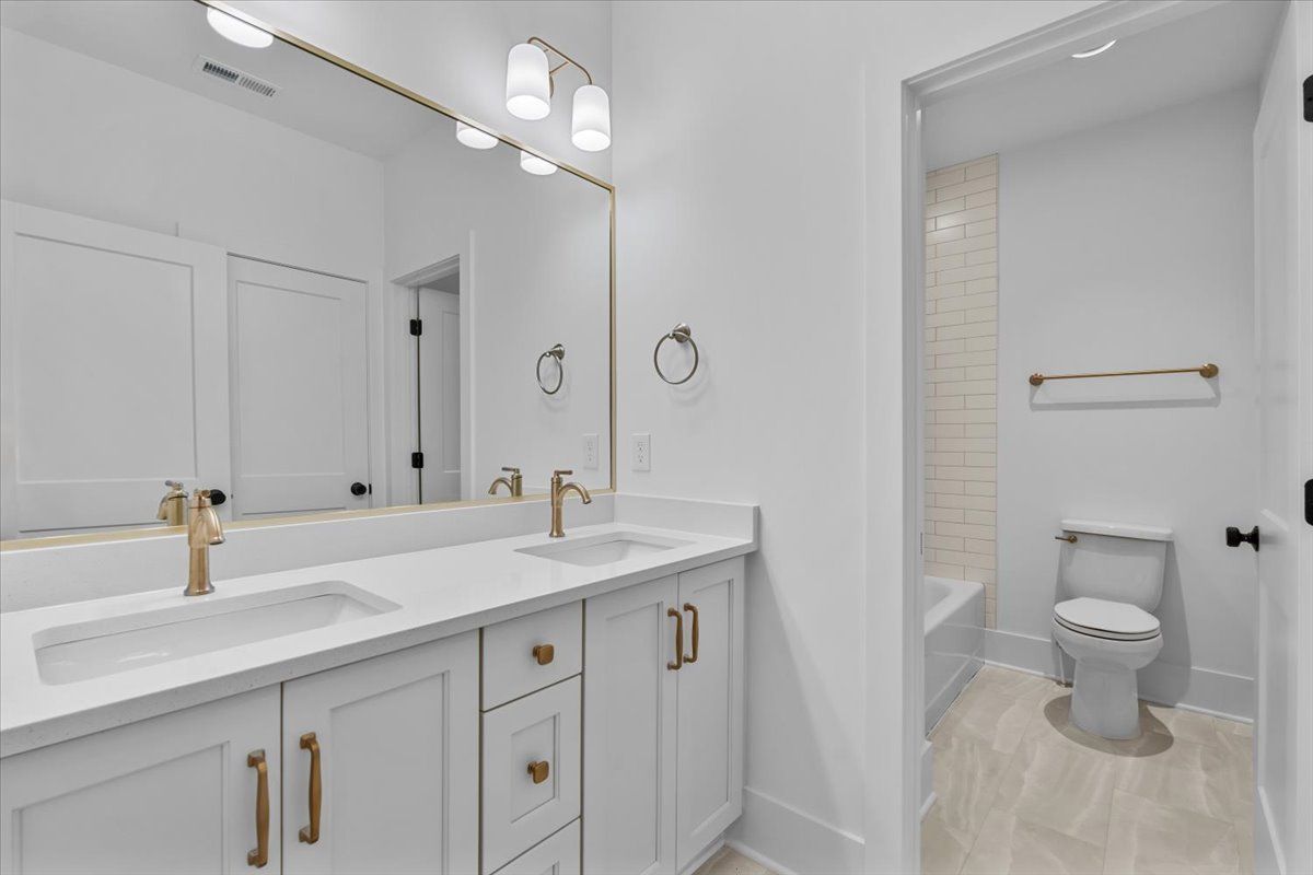 White bathroom with double vanity, gold fixtures, and a toilet visible in the doorway.