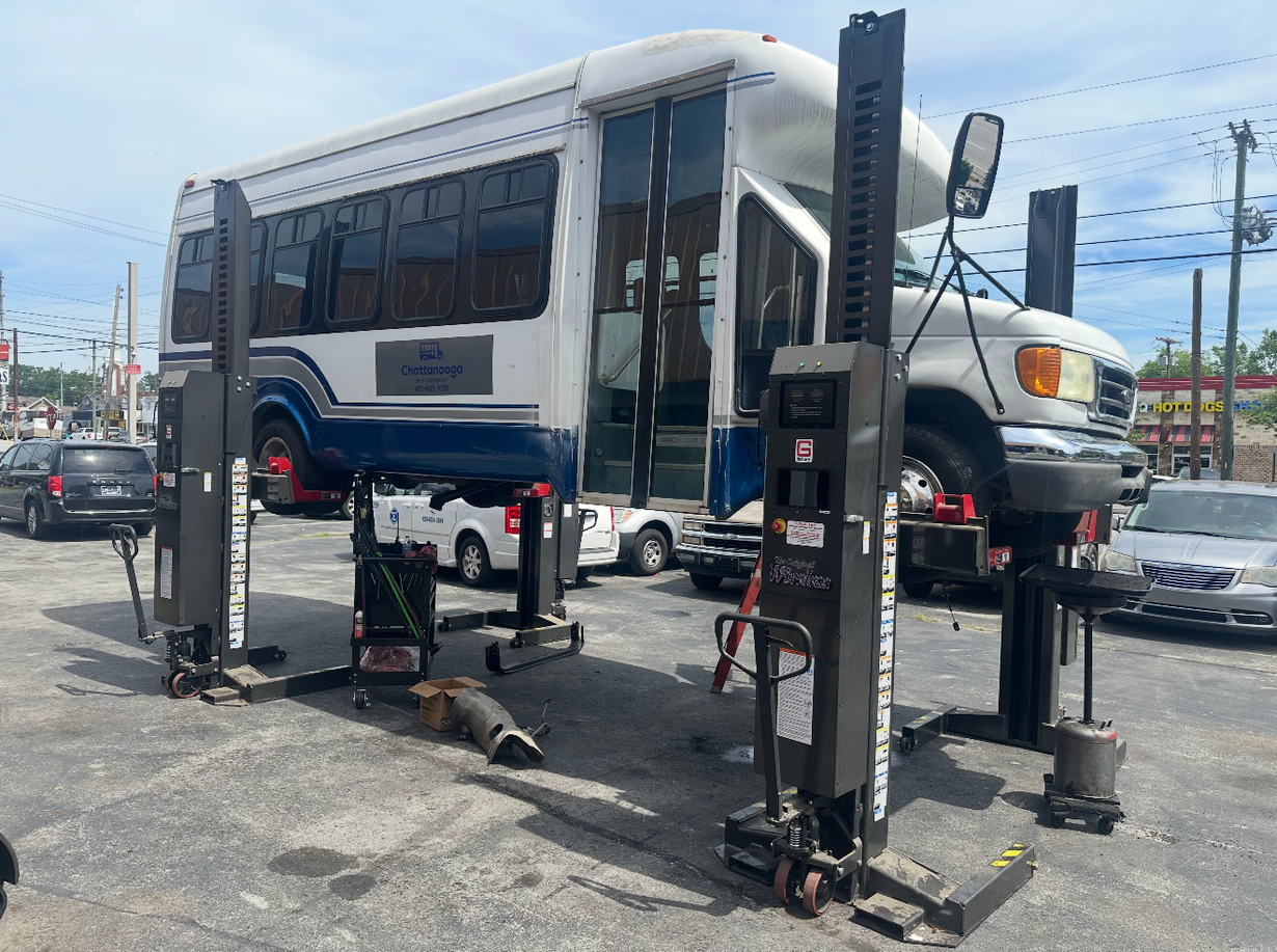 Bus lifted on a hydraulic jack in a parking lot, with its front wheels raised off the ground.