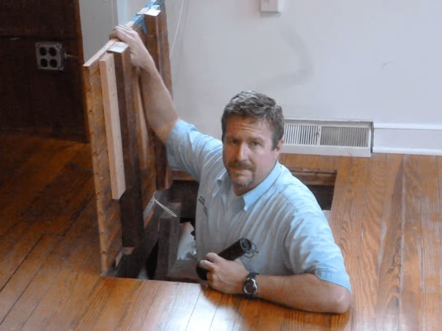 A person in a light blue shirt holds a flashlight while emerging from an open floor hatch in a room with wood flooring.