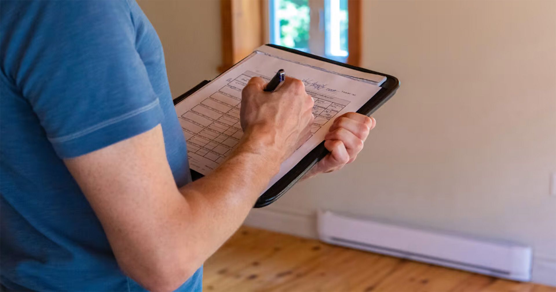 A person in a blue t-shirt writes on a clipboard inside a room with hardwood floors and a white baseboard heater.