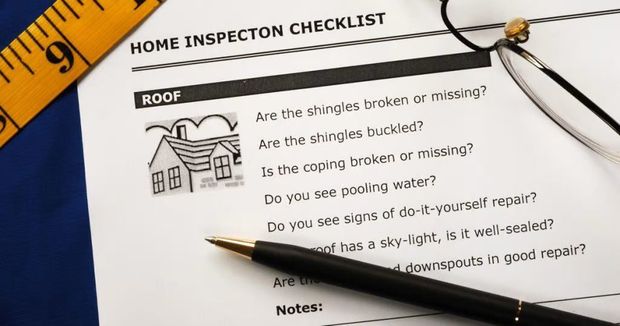 A close-up of a home inspection checklist, tape measure, and pen on a dark surface, focusing on the roof section.