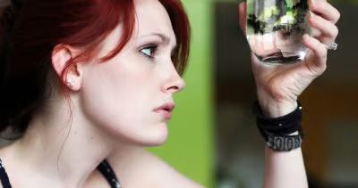 A person with red hair looking intently at a clear glass of water held up to their eye.