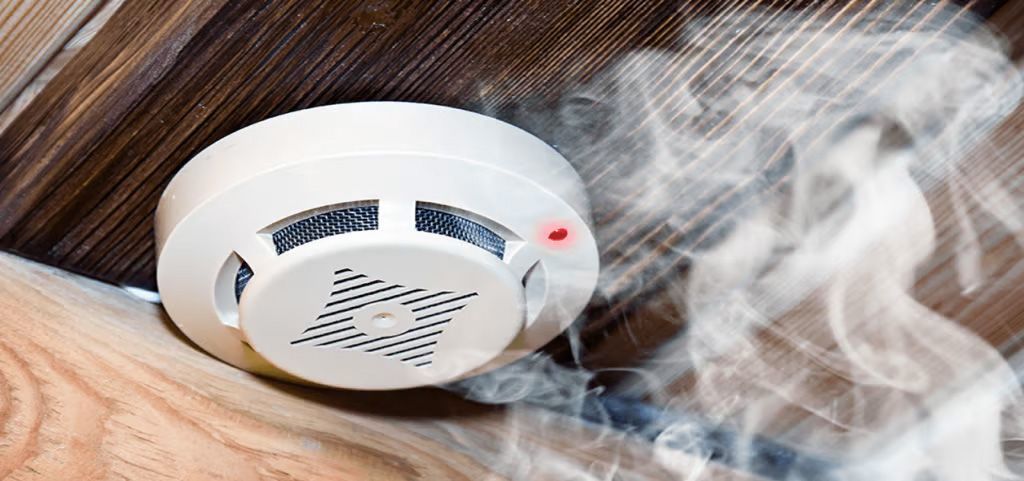 A white smoke detector mounted on a wooden ceiling with wisps of smoke nearby.