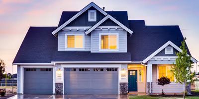 A two-story suburban house with blue siding, a dark roof, a two-car garage, and a teal front door at sunset.