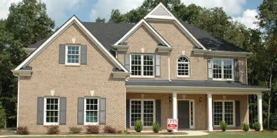 A two-story tan brick house with a dark shingled roof, grey shutters, a front porch, and a small yard with green trees.