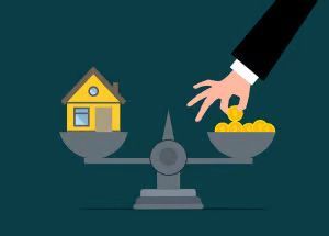 A hand placing gold coins on a balance scale opposite a yellow house, symbolizing the cost or value of real estate.