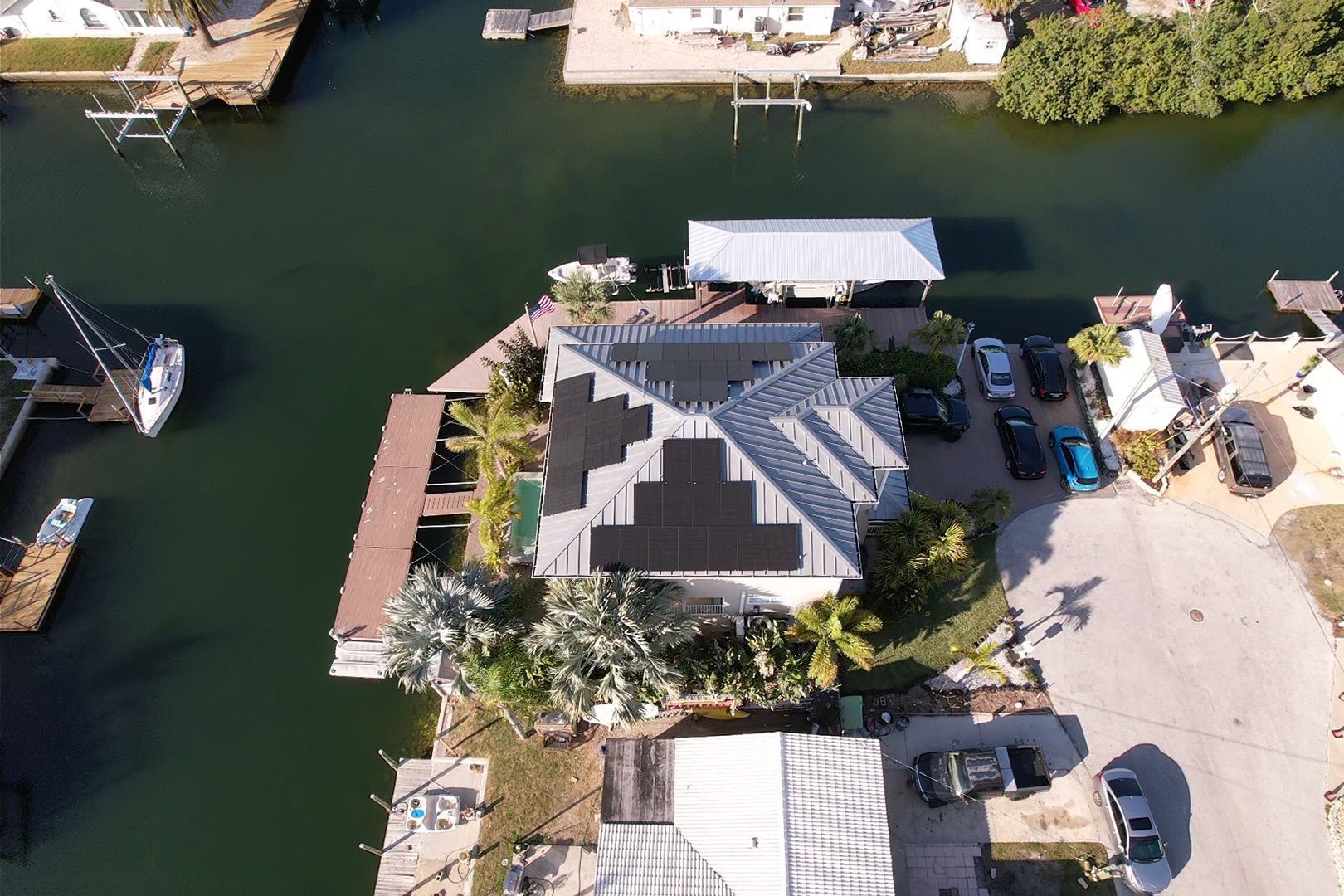 Aerial view of a canal-side house with a dark roof, dock, boats, and parked cars.