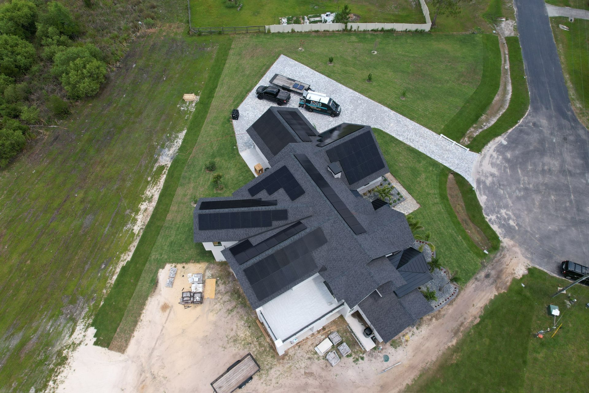 Aerial view of a dark-roofed house with driveway, lawn, and construction materials nearby.