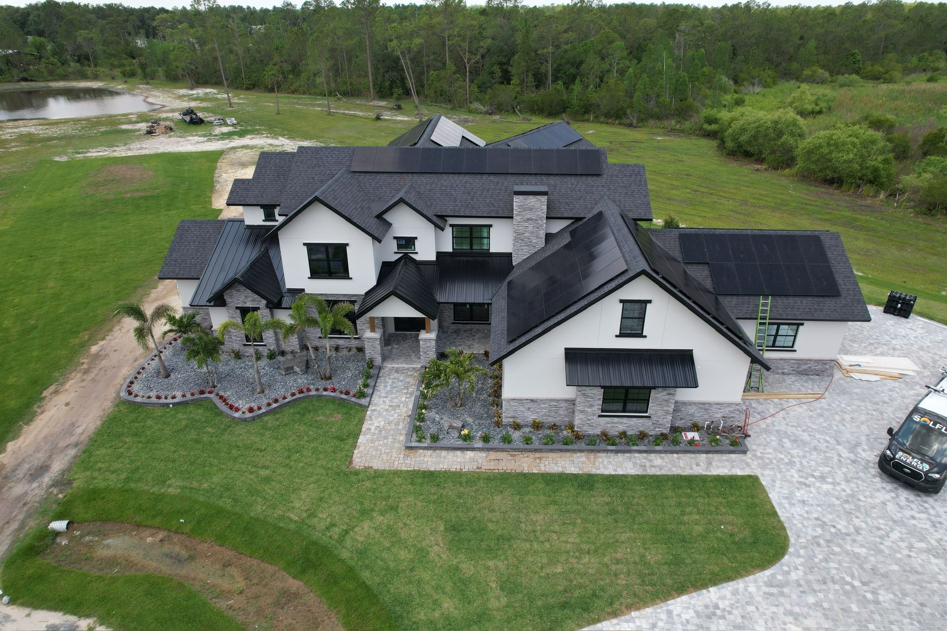 Aerial view of a large white-and-black house with a gravel driveway and wooded land around it.