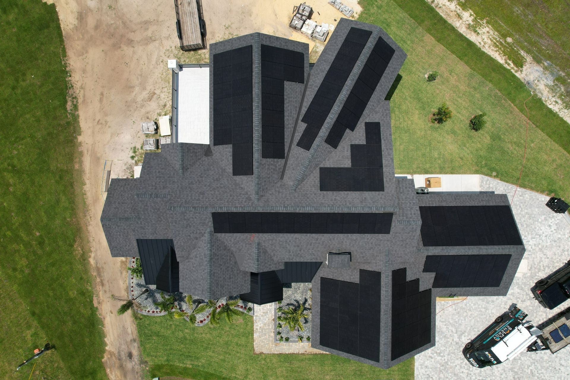 Aerial view of a dark-roofed house with multiple roof sections, surrounded by grass and a driveway.