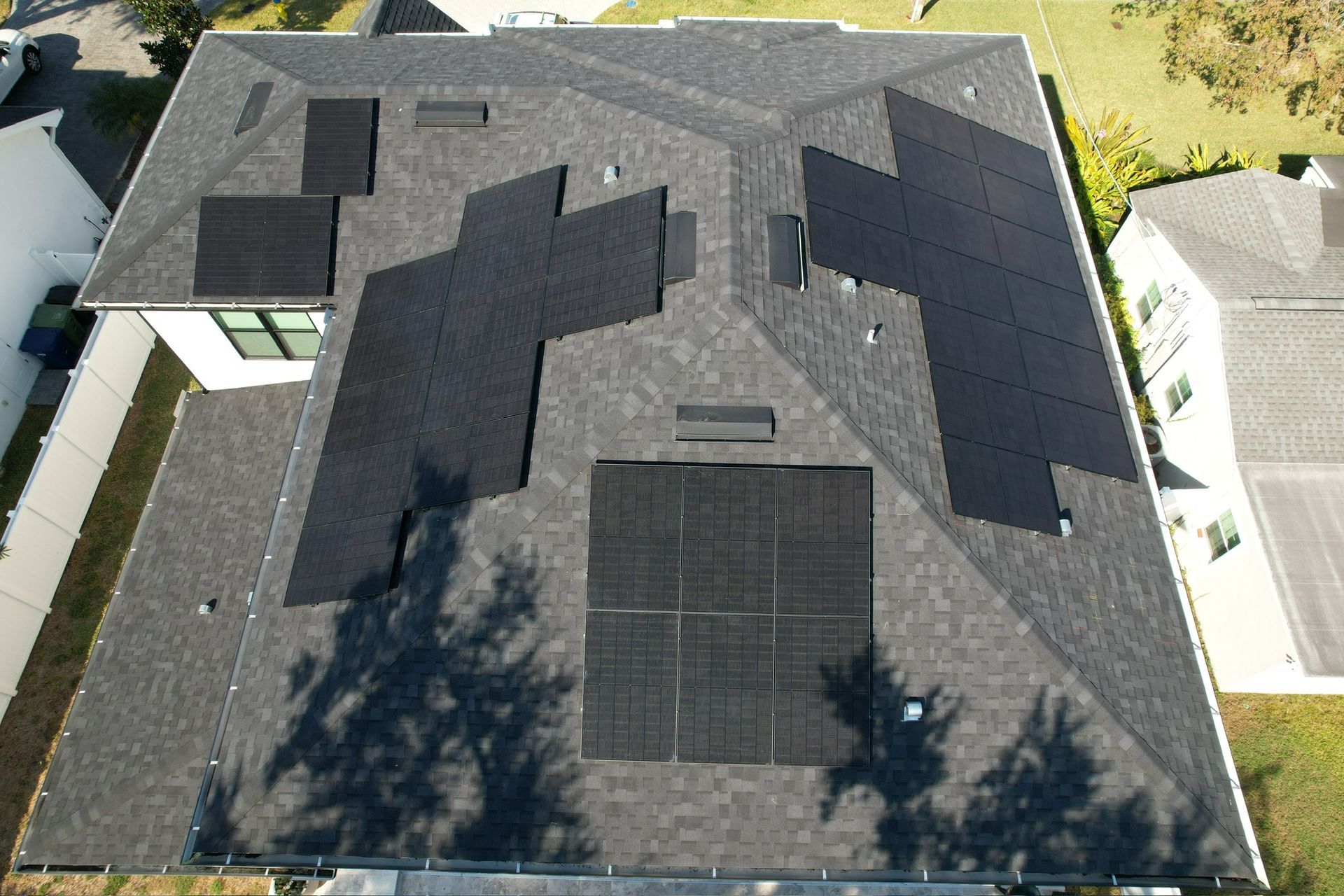 Aerial view of a house roof with dark shingles and a solar panel, surrounded by trees and neighboring homes.