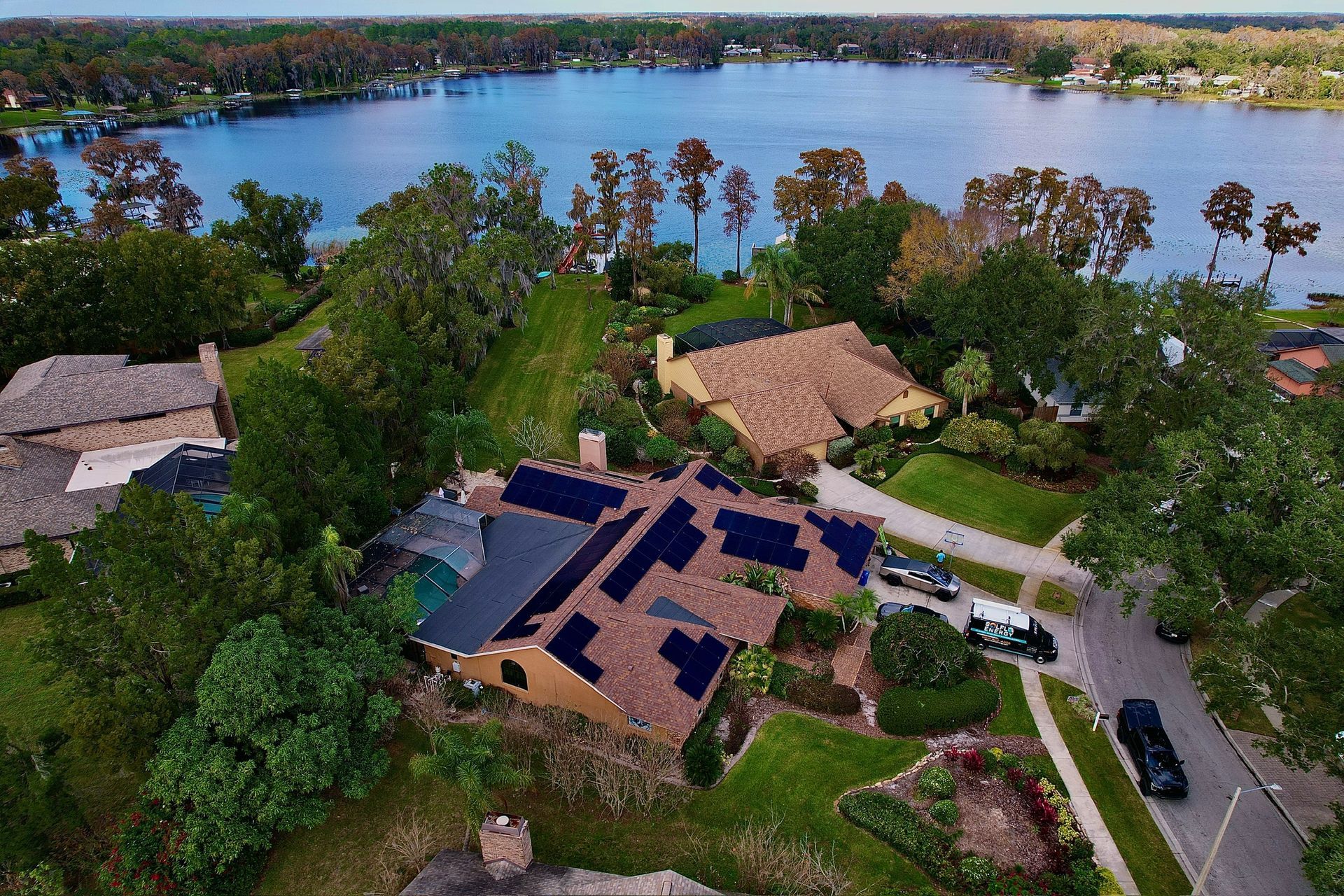 Aerial view of a lakeside home with dark solar panels, surrounded by trees and a winding road.