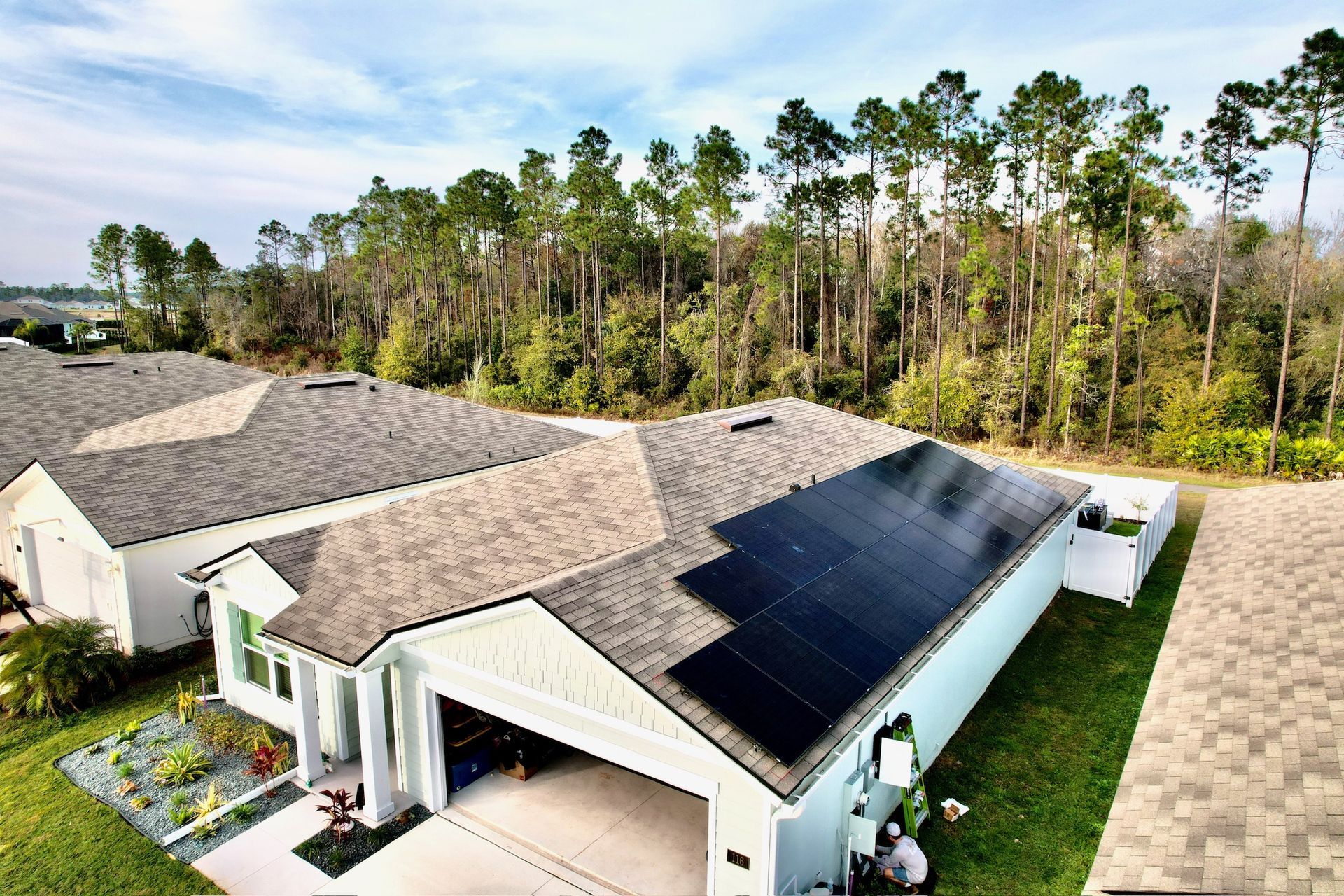 Aerial view of a suburban home with solar panels on the roof beside a wooded area.
