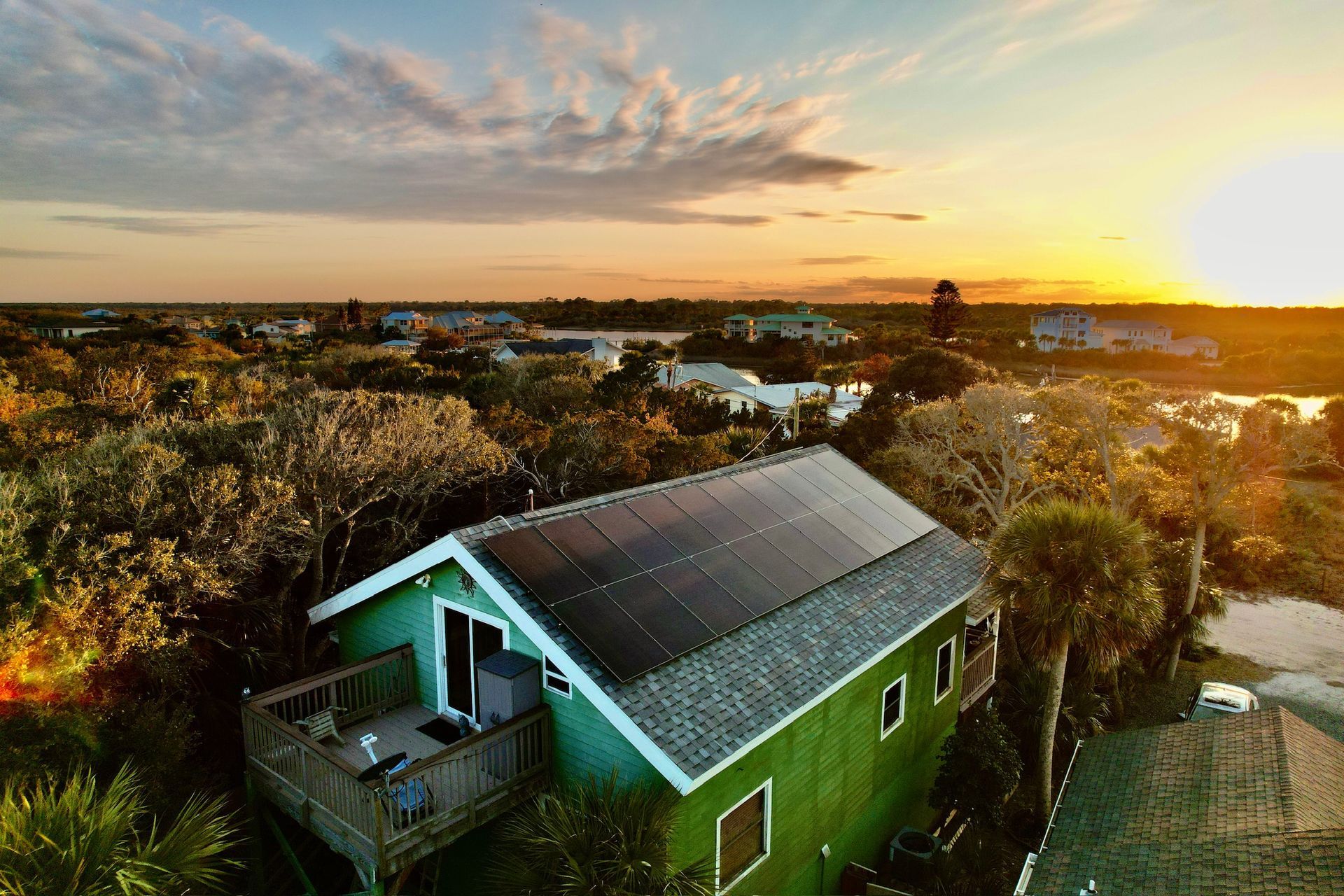 Green house with solar panels at sunset, overlooking a tree-filled neighborhood and glowing sky.