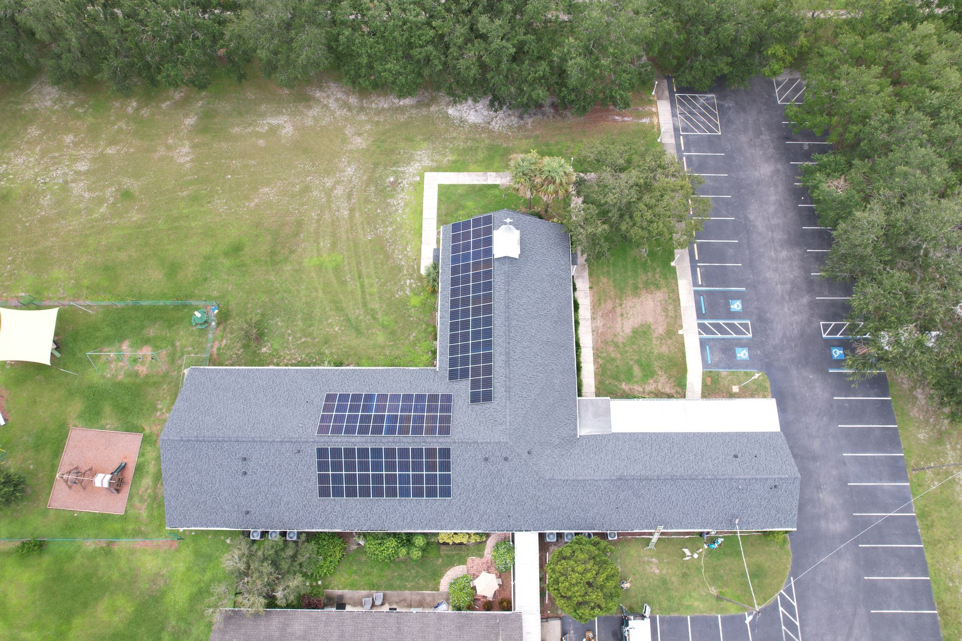 Aerial view of a gray-roofed building with solar panels beside a parking lot and grassy area.