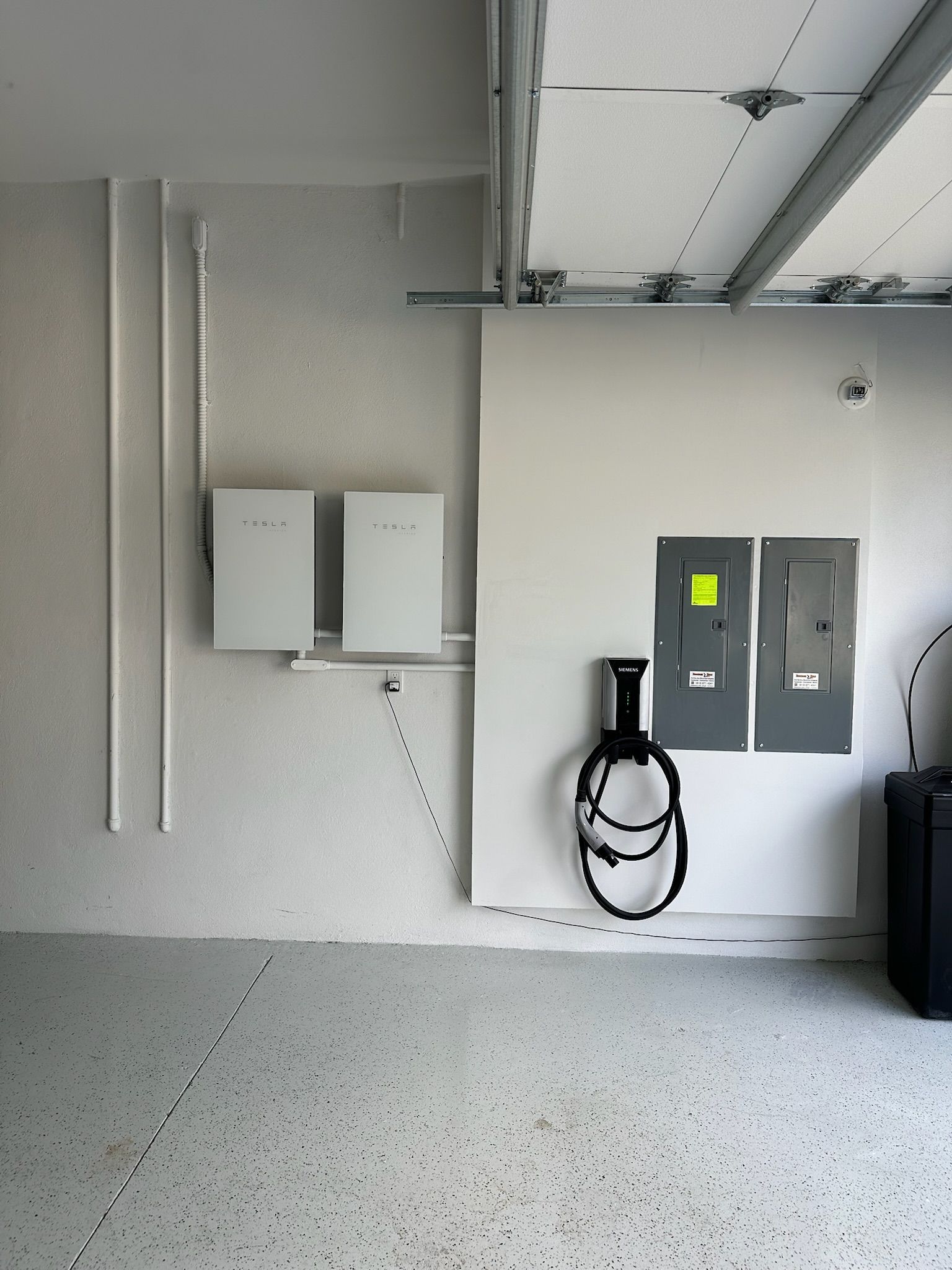 Garage wall with utility boxes, conduit, and a coiled black cable on a gray speckled floor.