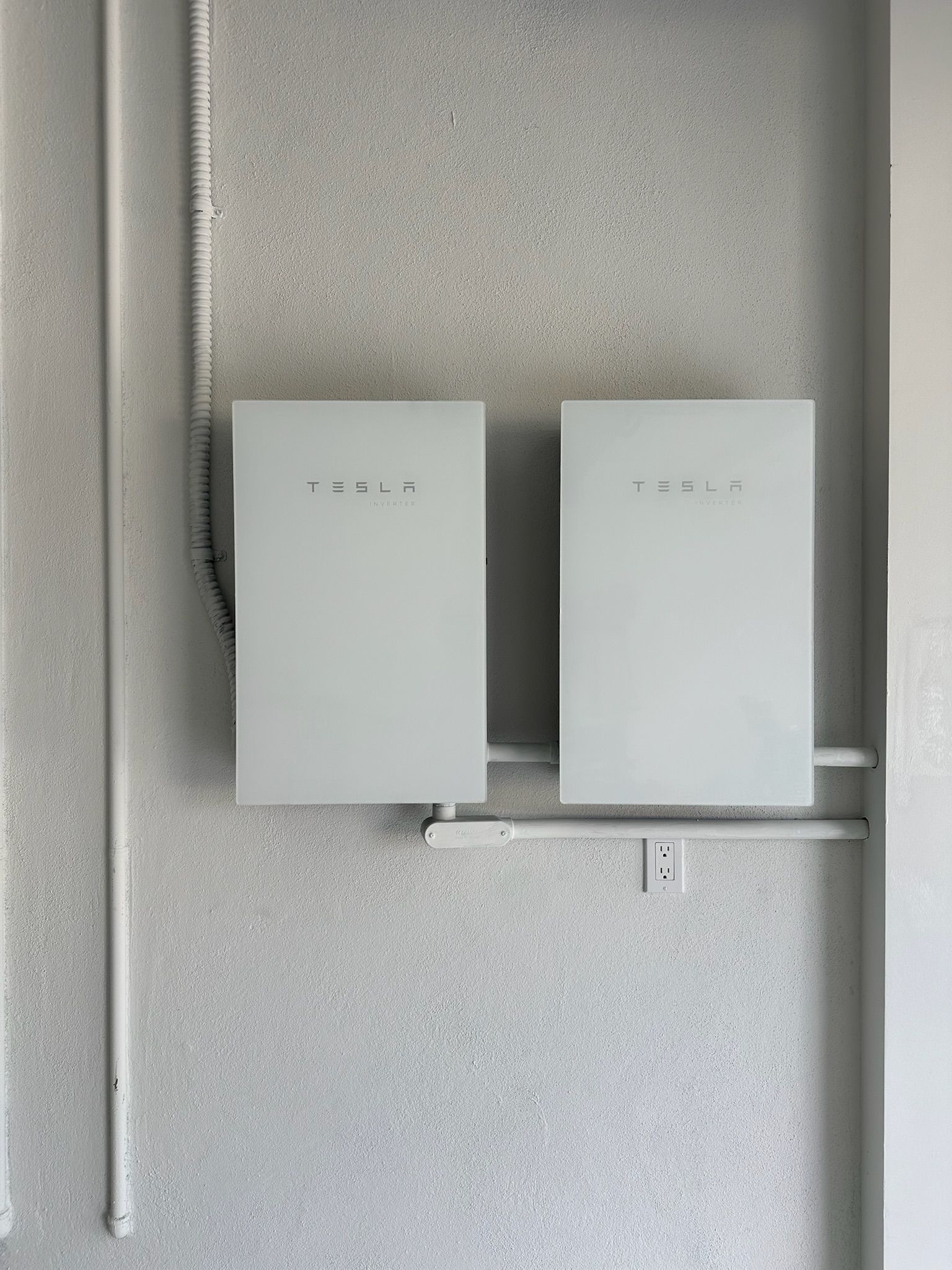 Two white utility boxes mounted on a gray wall with exposed pipes and conduit.