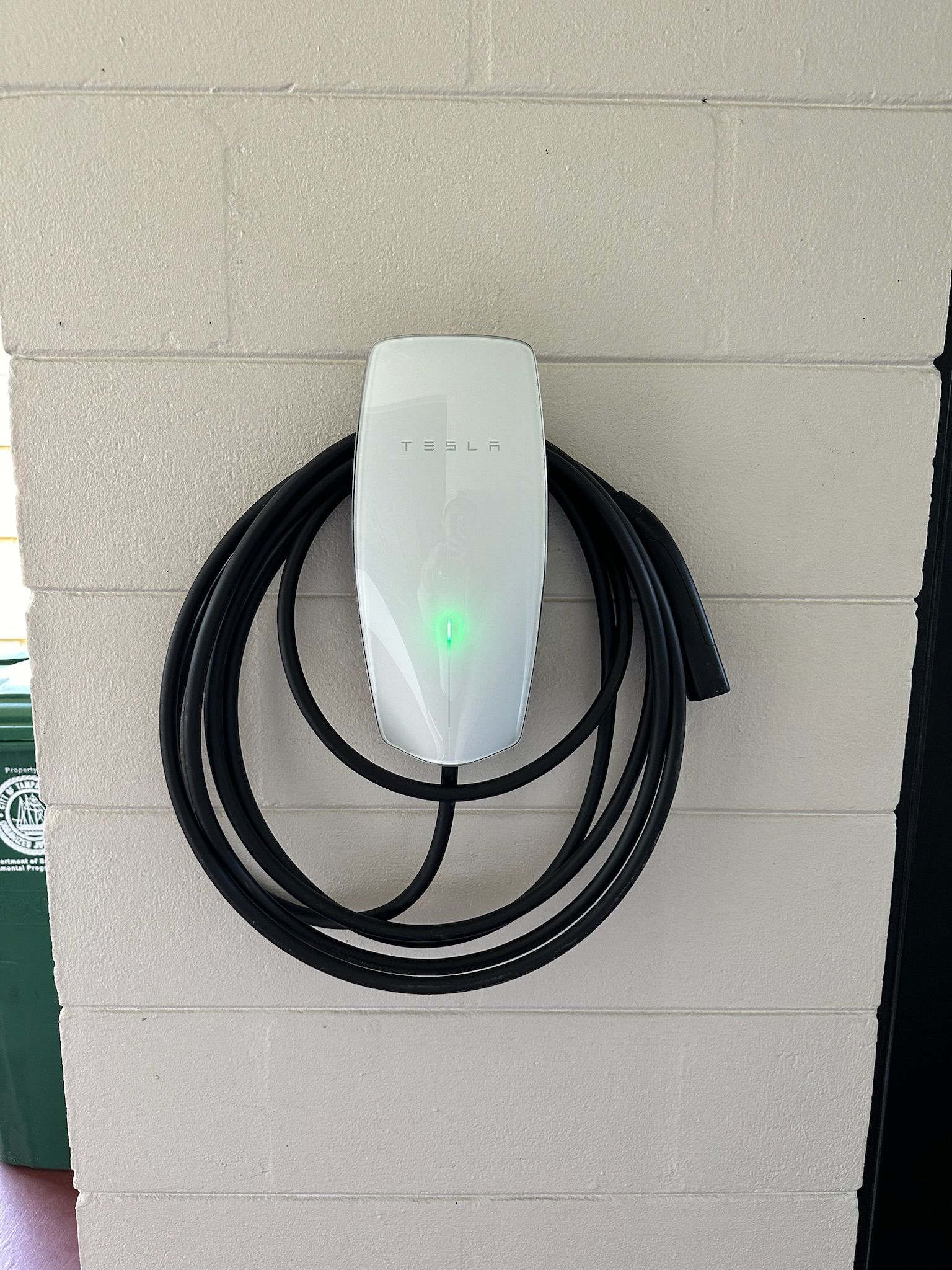 Wall-mounted white EV charger with coiled black cable on a gray wall, green light glowing.