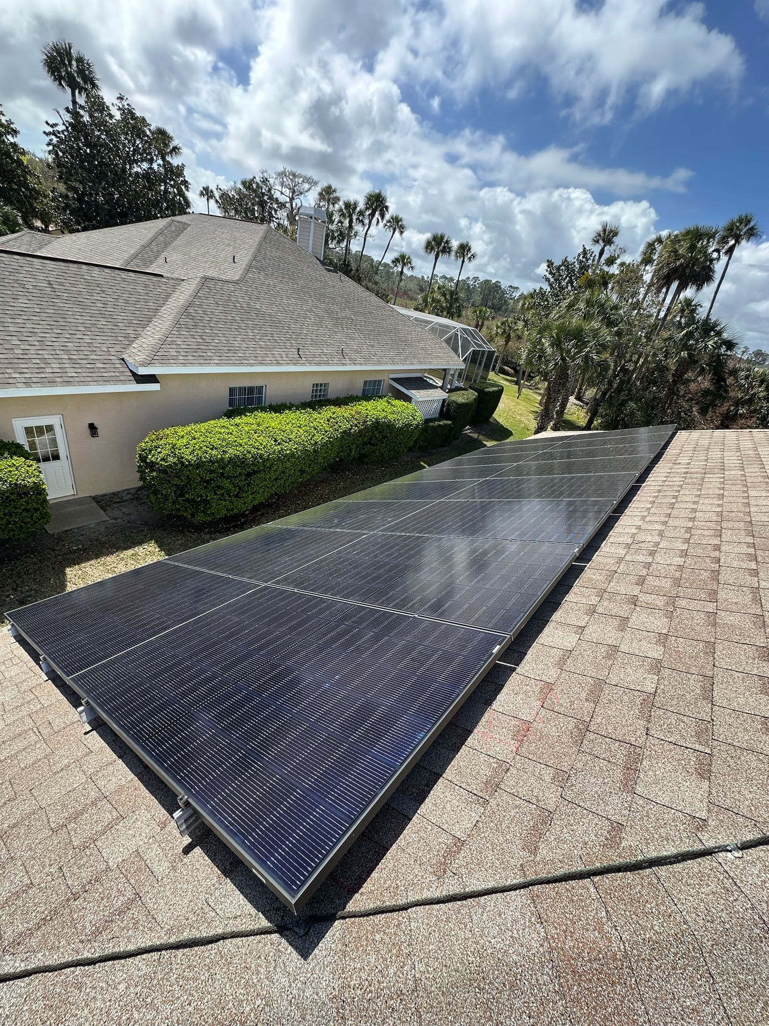 Solar panel arrays on a rooftop beside a house under a partly cloudy sky.