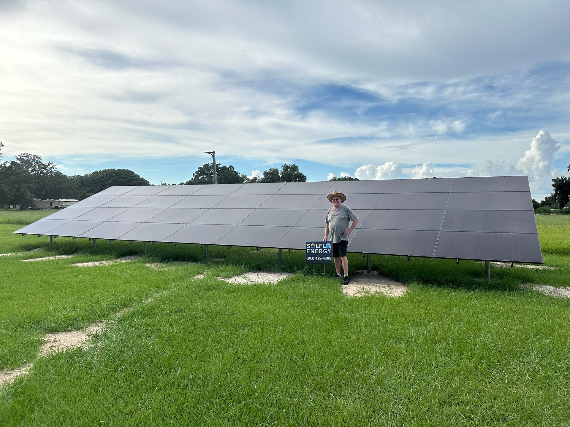 Person standing beside a large solar panel array on a grassy field under a cloudy sky.