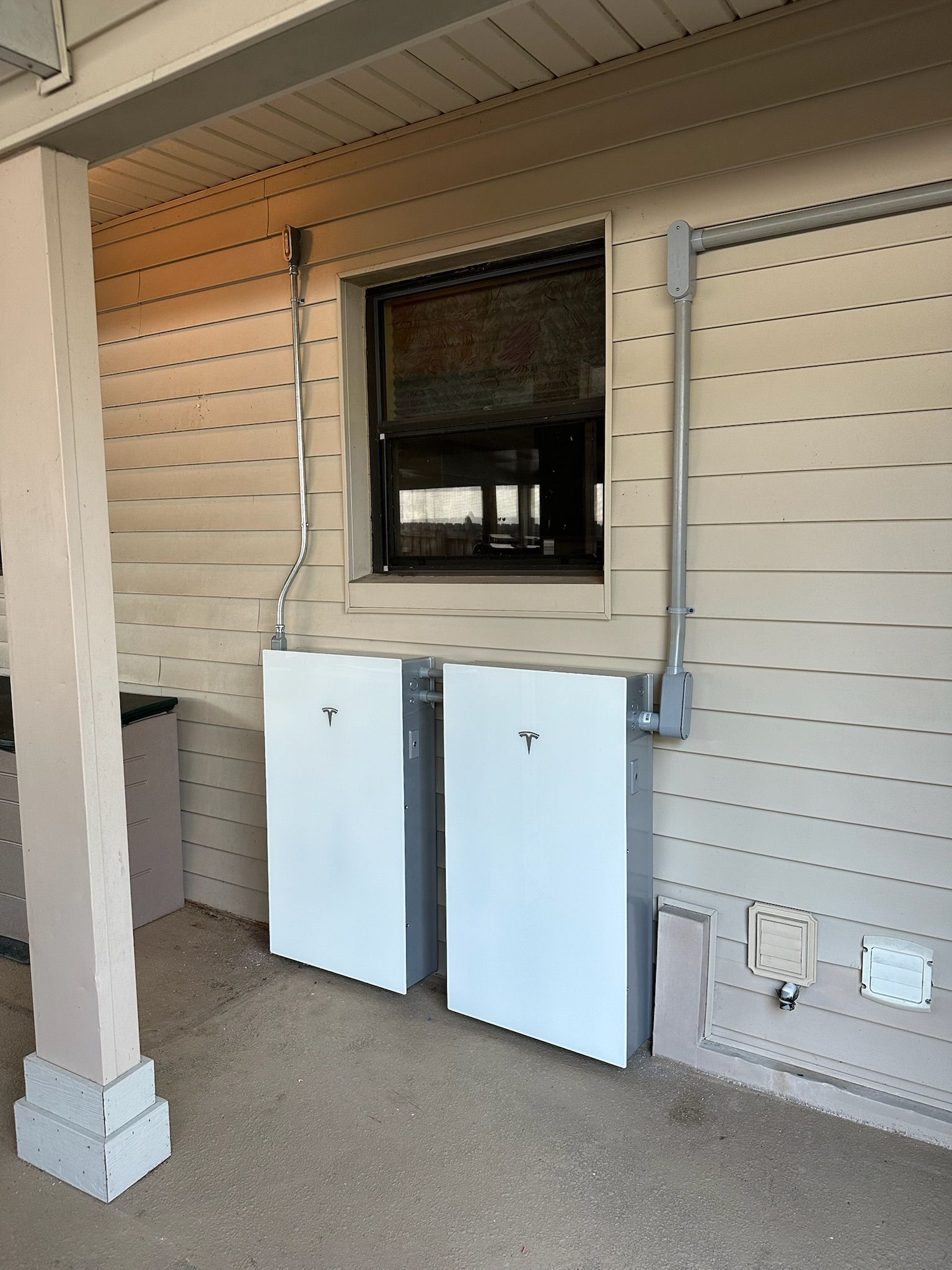Two white utility cabinets beneath a window on a covered porch, with wall-mounted conduit and outlets.