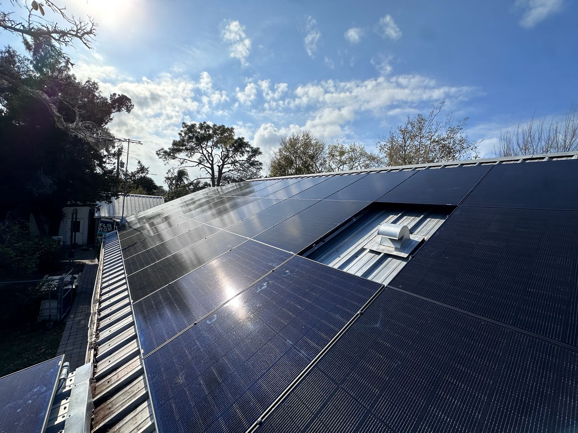 Solar panels on a sloped roof under a bright blue sky with clouds and trees nearby.