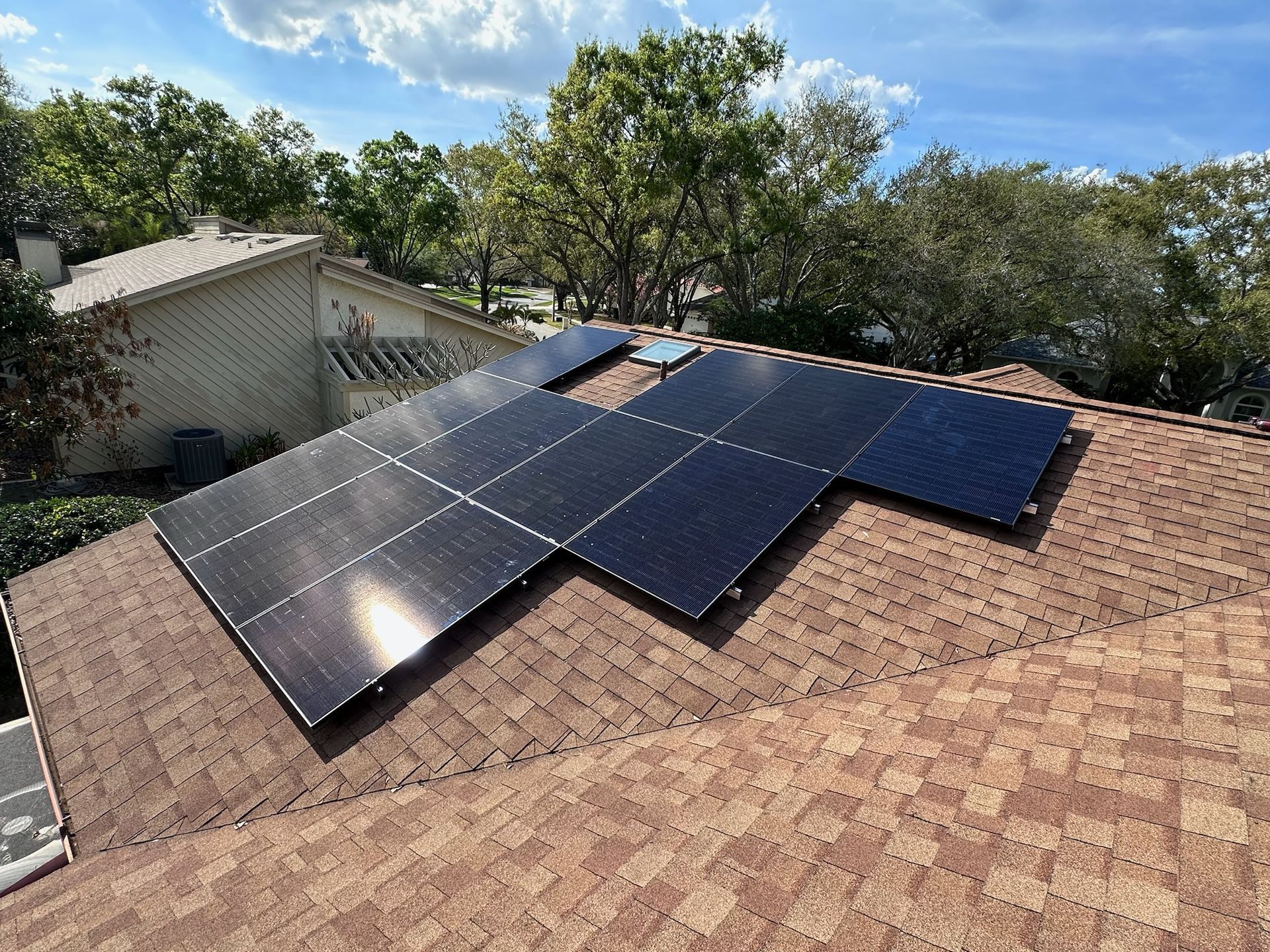 Solar panels on a shingled roof under a blue sky, with trees in the background.