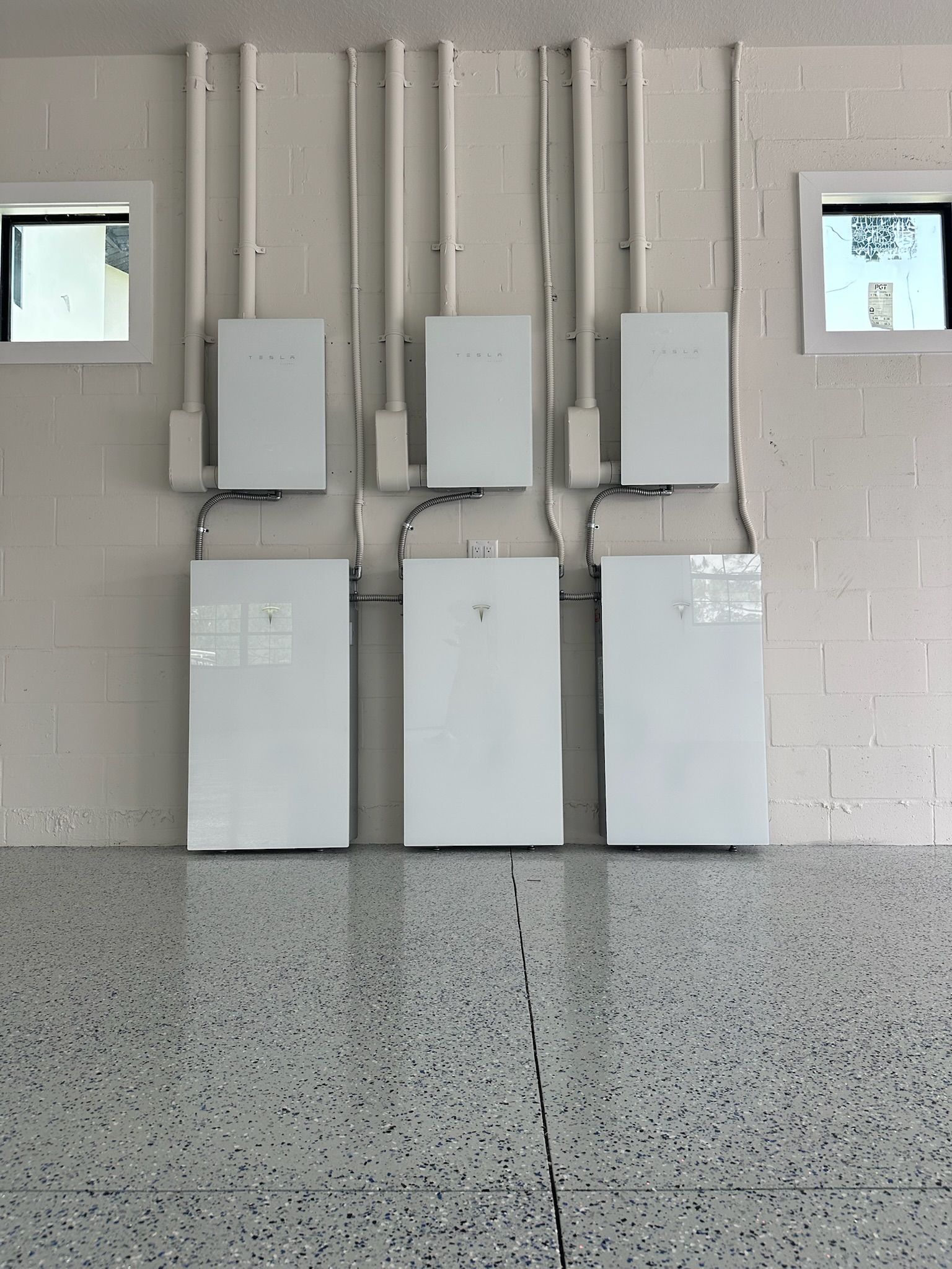 Wall-mounted utility panels and conduits in a garage-like room with a speckled floor and small windows.