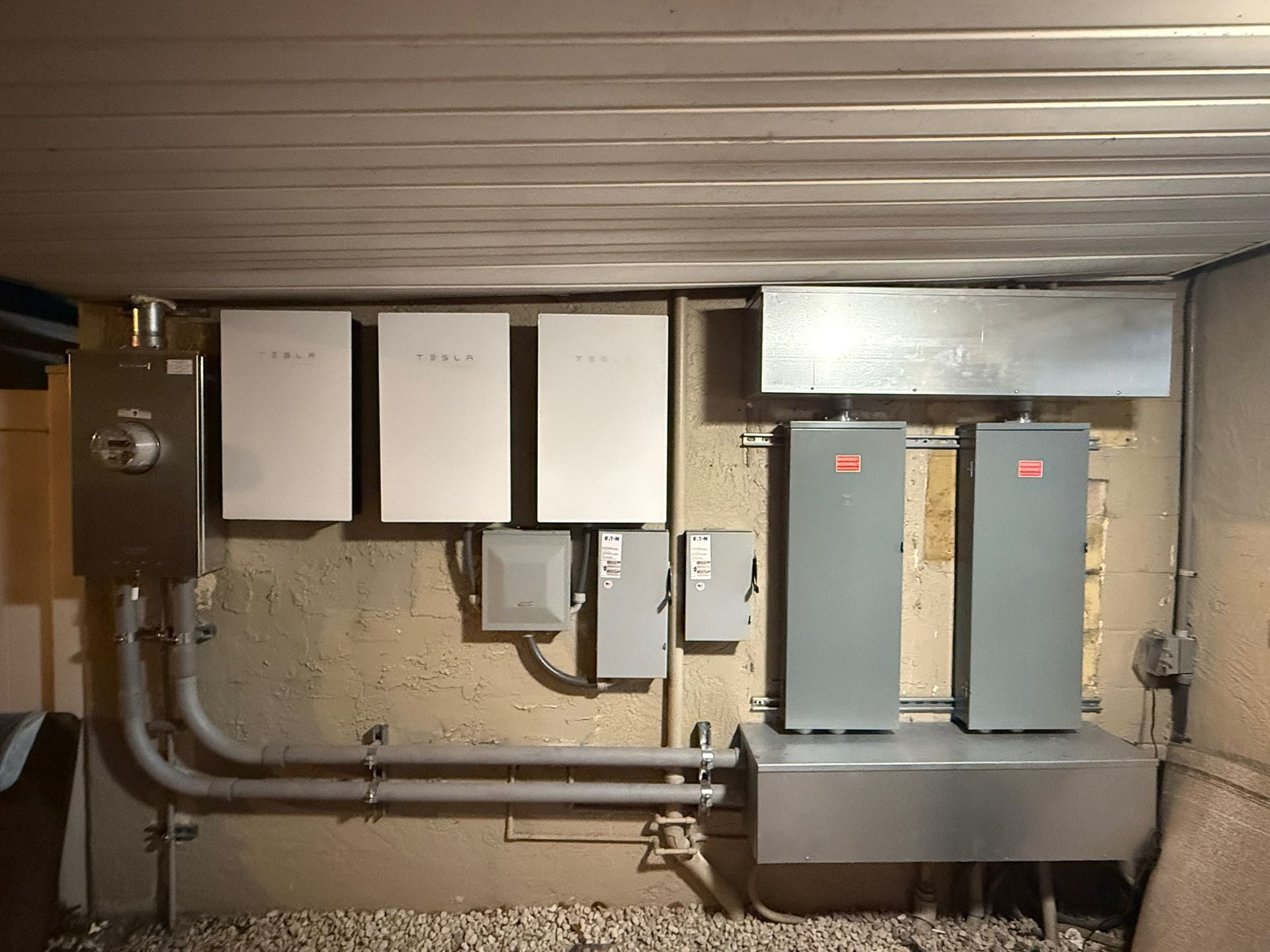 Utility room with electrical panels, gray equipment boxes, and exposed conduit along a concrete wall.