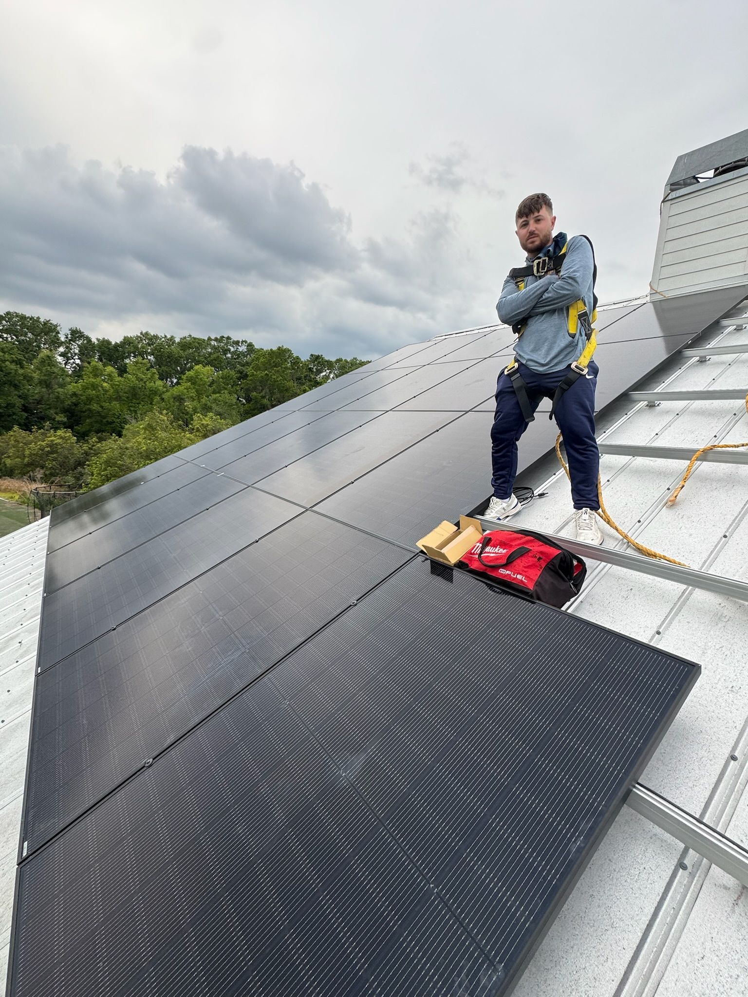 Worker on a steep metal roof installing dark solar panels under cloudy skies.