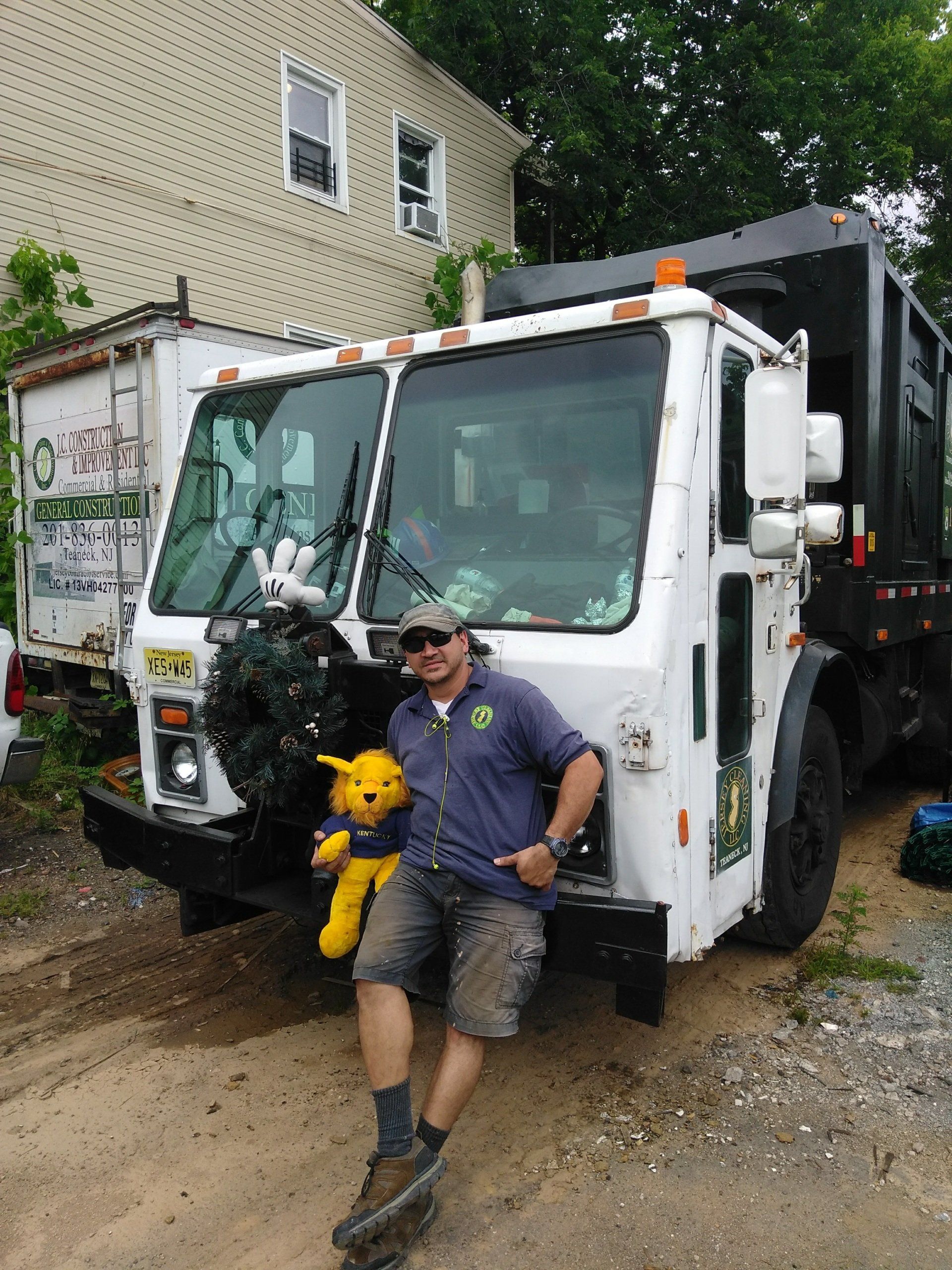 A man is standing in front of a Jersey Cleaning garbage truck holding a stuffed animal.