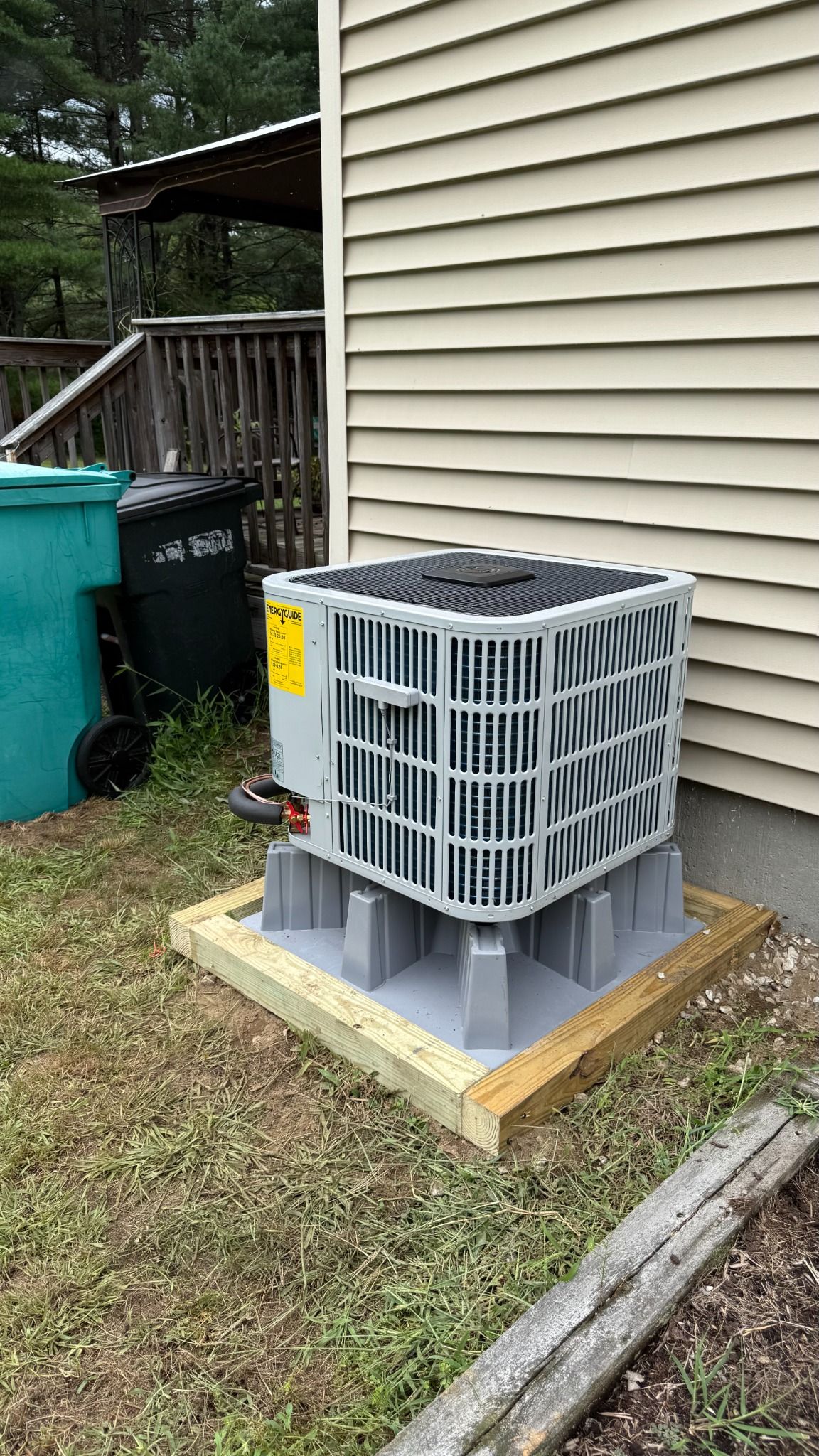 Outdoor air conditioner unit on a wooden platform beside a house, next to trash bins and grass.