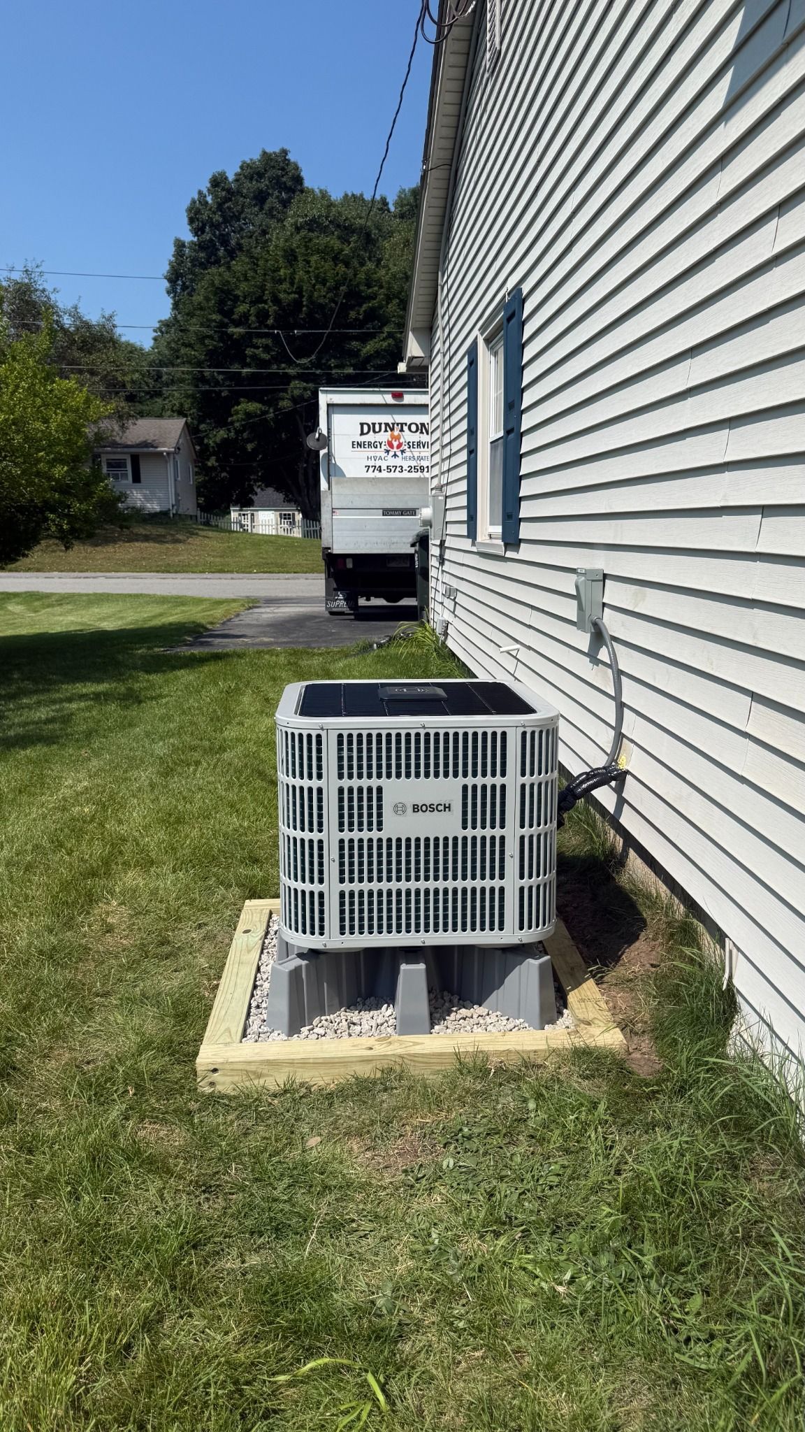 Outdoor AC unit beside a white house, with a service truck visible in the driveway.