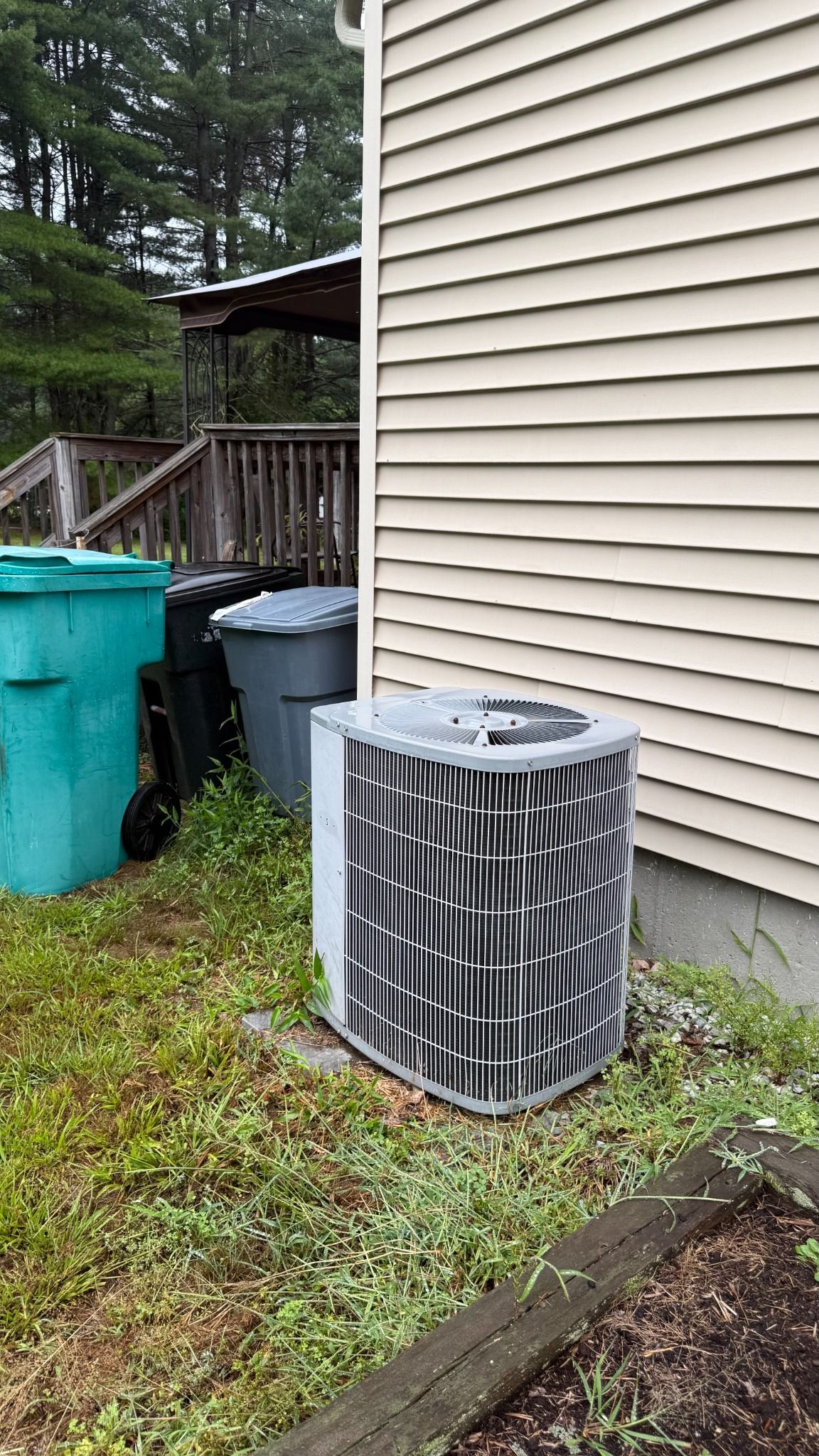 Outdoor air conditioner unit beside beige house wall, near trash bins and a wooden deck in a grassy yard