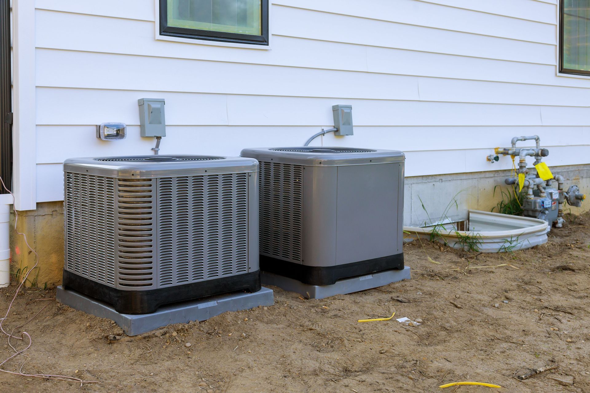 Two outdoor air conditioning units beside a white house wall on bare dirt.