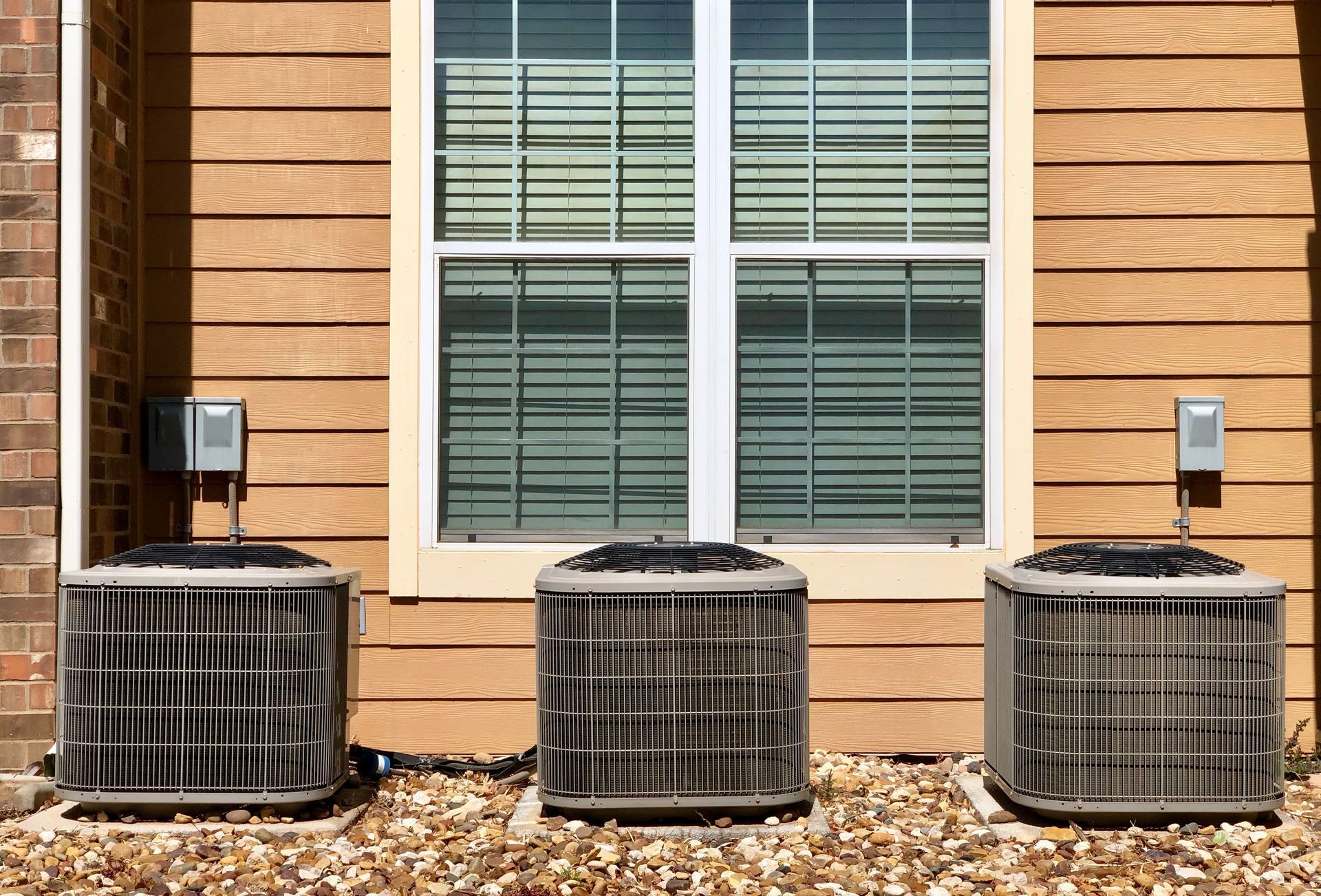 Three outdoor HVAC units under a window on a wood-sided house with mulch below.