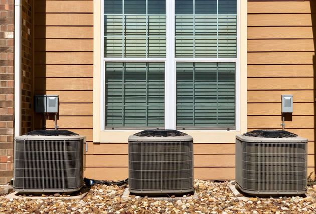 Three outdoor HVAC units under a window on a wood-sided house with mulch below.