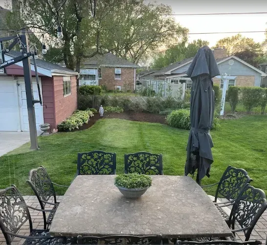 Backyard patio with metal table, chairs, closed umbrella, and a neatly landscaped lawn and garden.