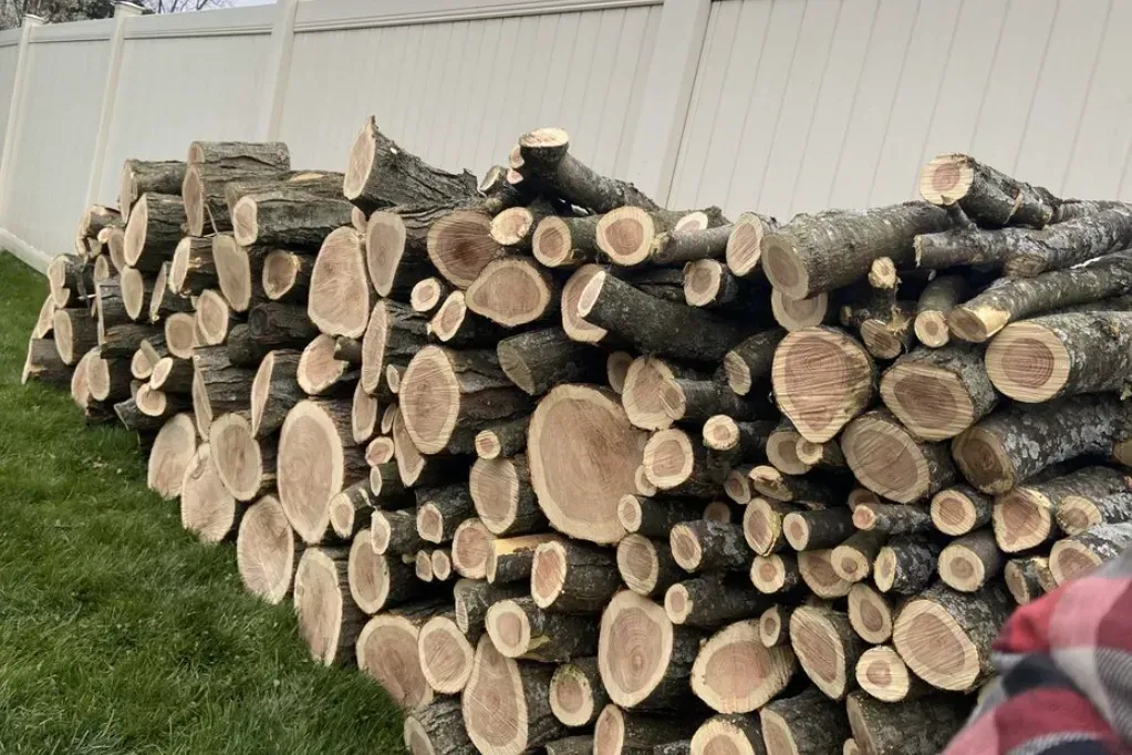 Stacked cut firewood logs beside a white building, with grass in the foreground.