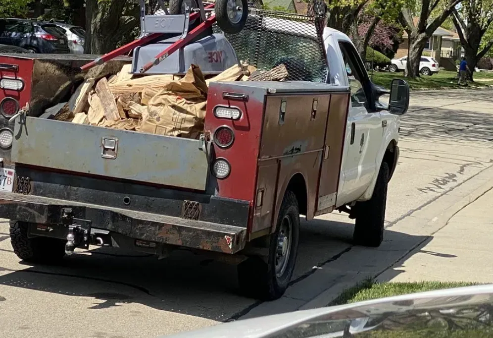 White pickup truck with a utility bed loaded with large bags, parked on a tree-lined street.