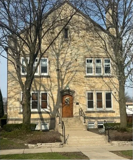 Tan stone apartment building with steep roof, central wooden door, and leafless trees in front