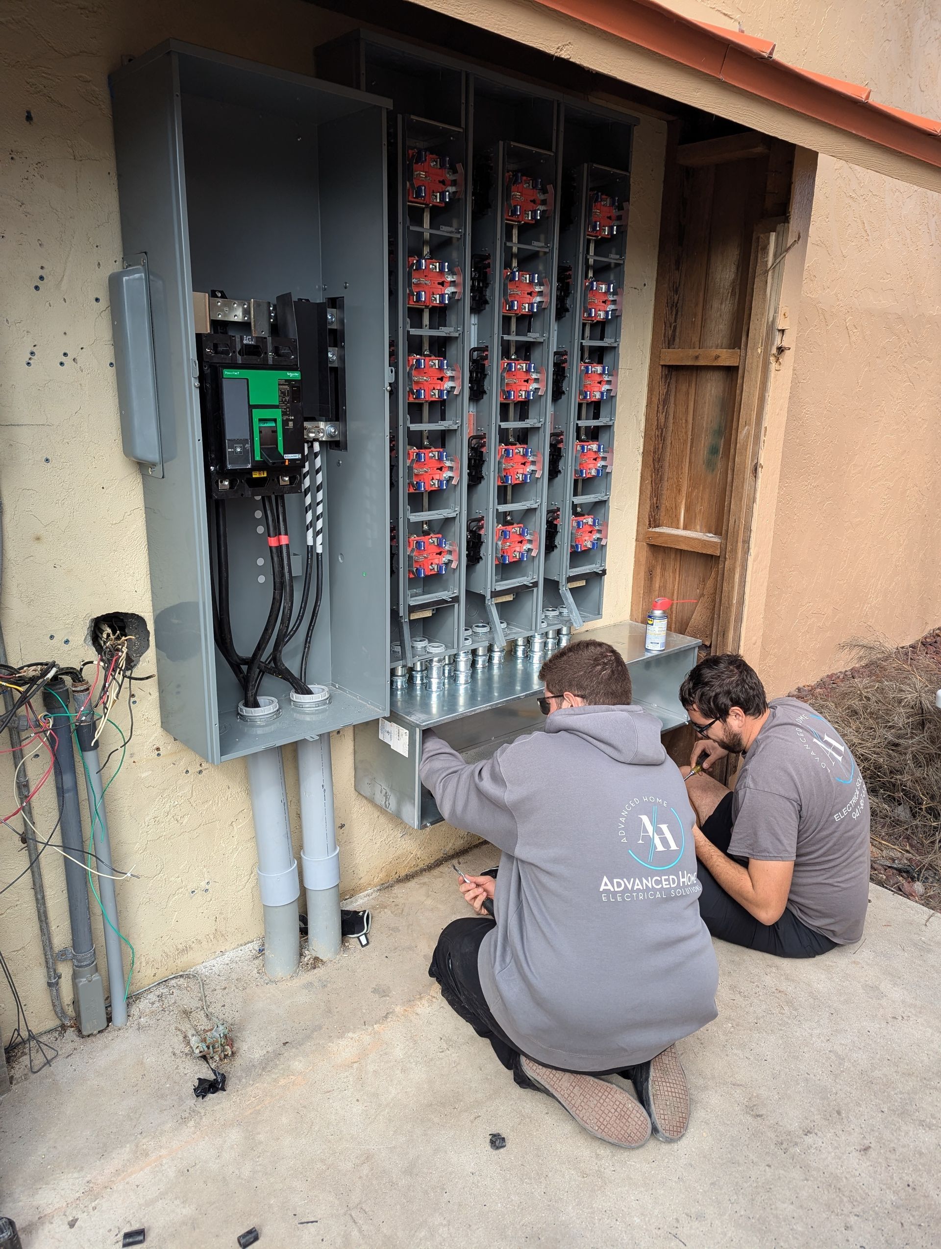 Two workers kneel beside an open electrical cabinet with wiring and circuit panels outdoors.