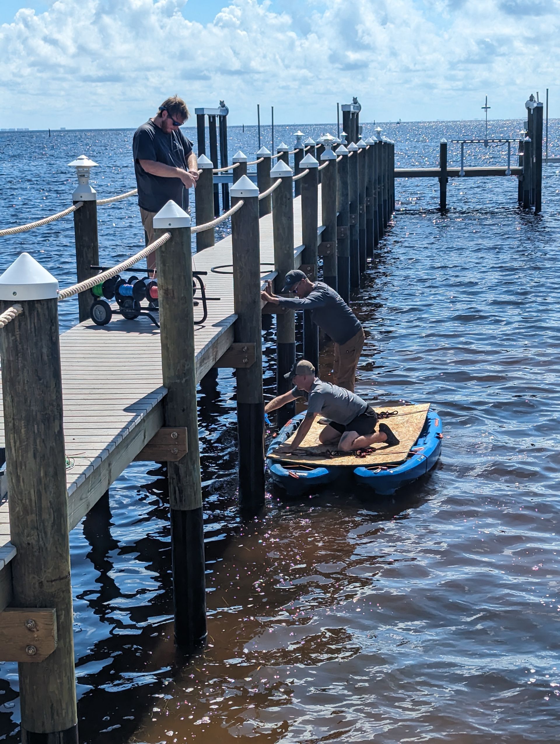 People loading a paddleboard at a wooden dock on a sunny waterfront.
