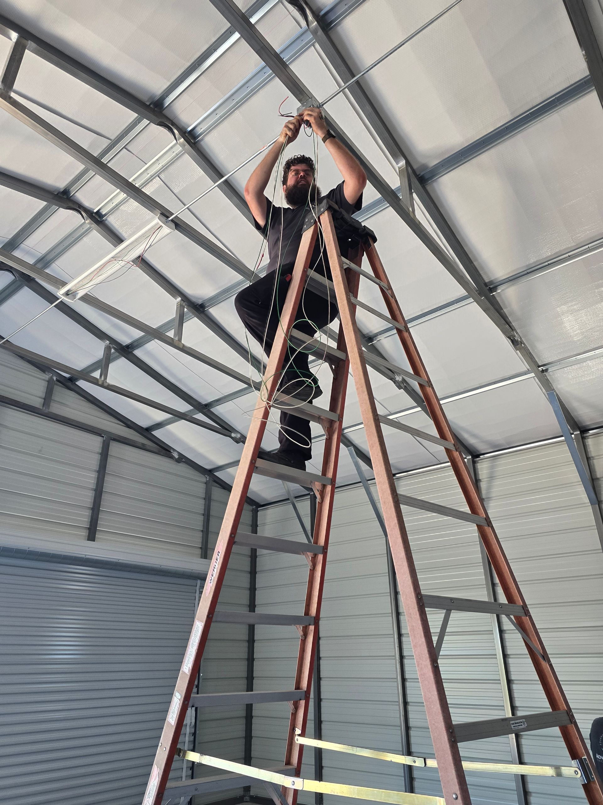 Person on a tall ladder working near the ceiling inside a metal-roofed building.