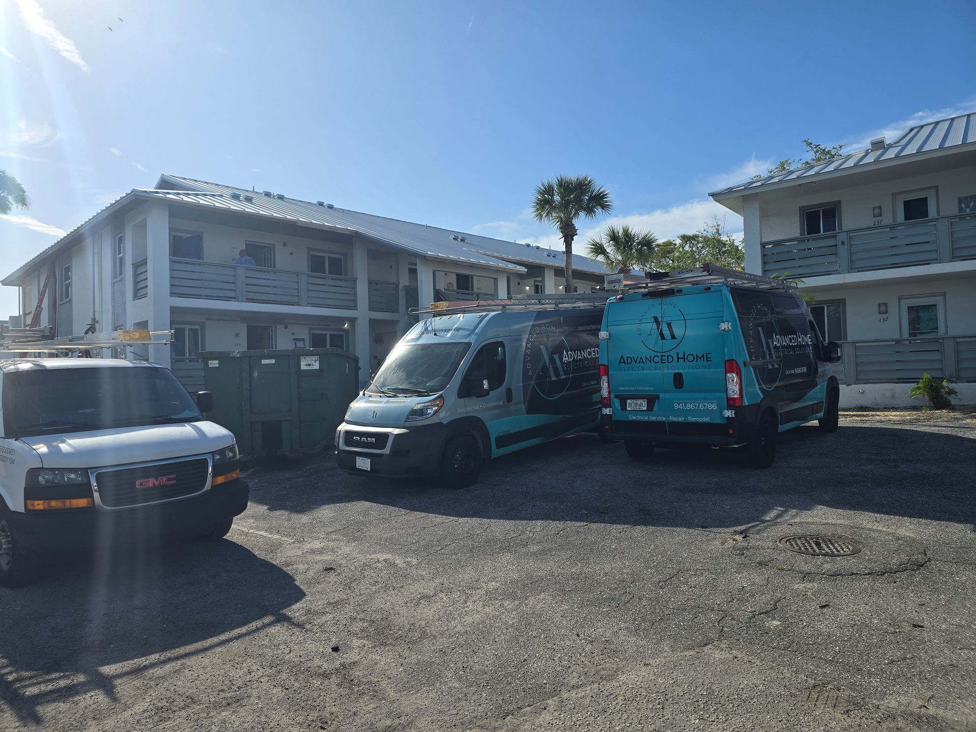 Two white vans parked in a gravel lot beside two-story apartment buildings under a blue sky.