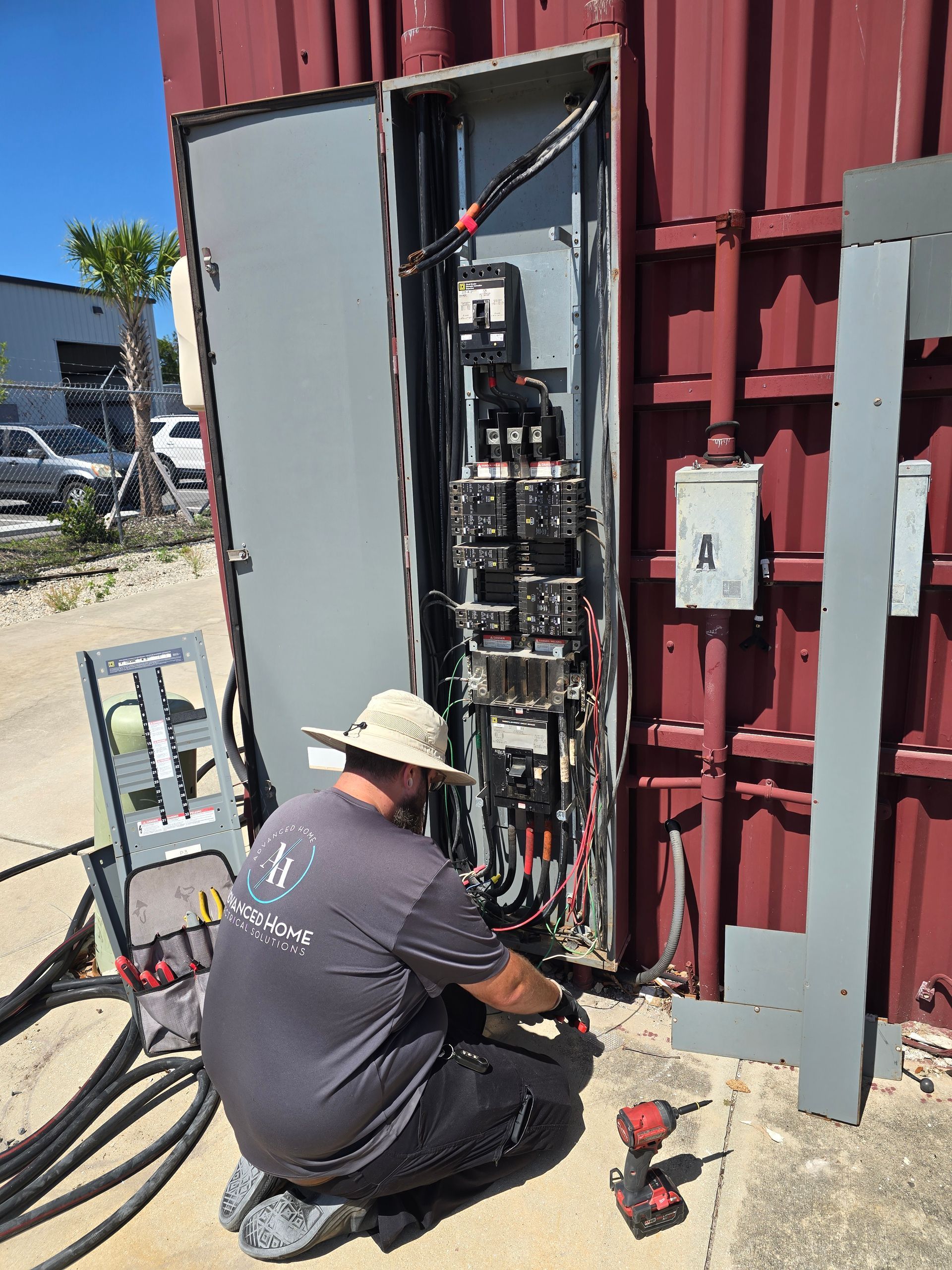 Technician working on an open electrical panel outside a red building, with tools and cables nearby.