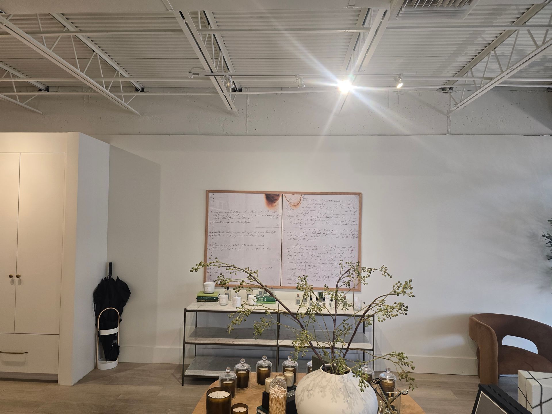 Minimalist white room with shelving, plants, wall art, and a woven chair under exposed ceiling beams.